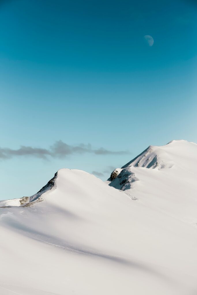 Serene view of snowy peaks in Switzerland under a clear blue sky with the moon visible.
